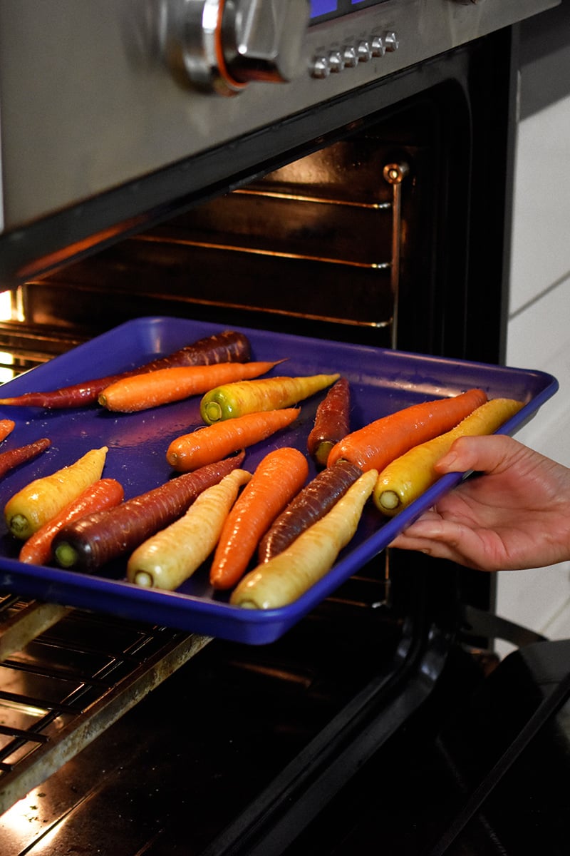 Someone putting a blue rimmed baking sheet filled with rainbow colored carrots into an open oven.