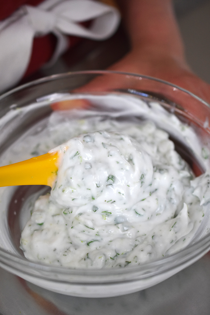 A closeup of the herby coconut yogurt being mixed in a clear glass bowl with a small yellow spatula.