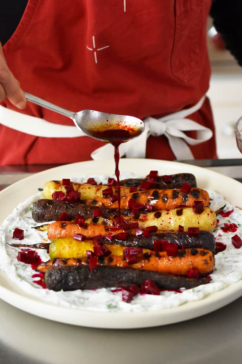 Someone in a red apron is adding beet vinaigrette on top of grilled carrots on coconut yogurt.