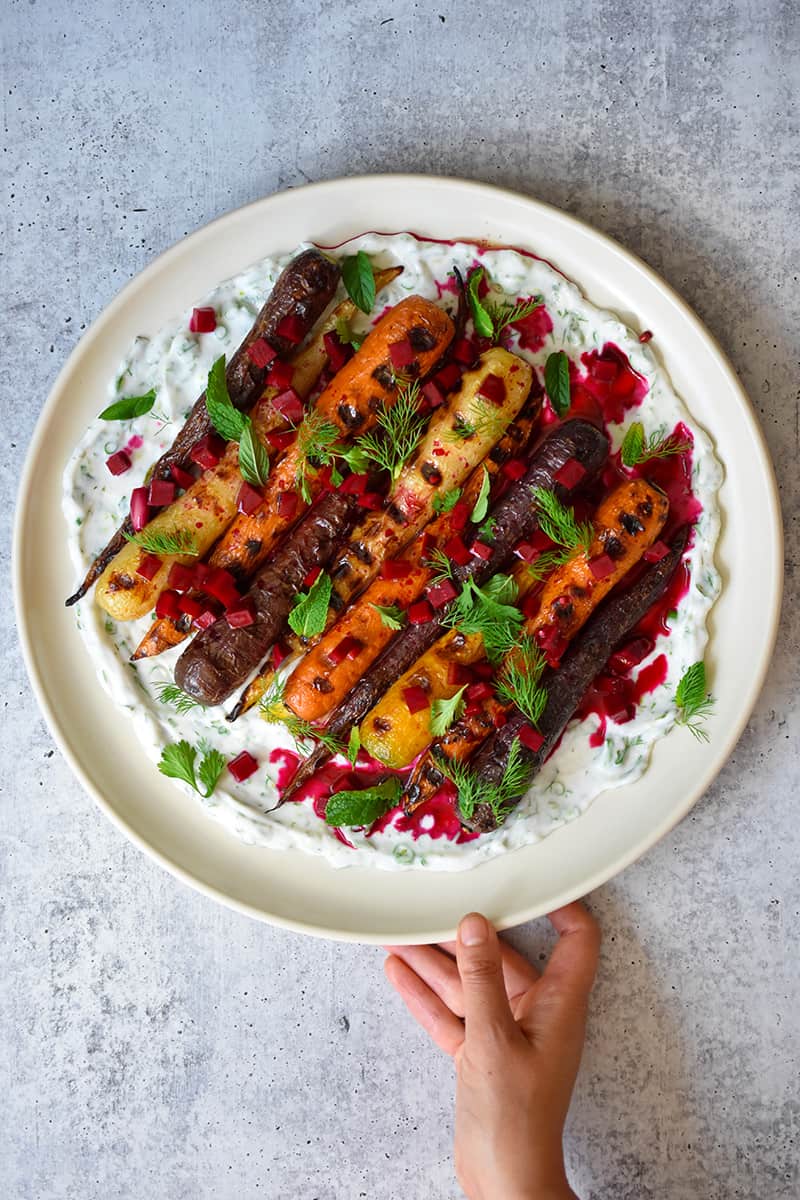 A hand is holding a round platter filled with grilled carrots with herby coconut yogurt and spicy beet vinaigrette