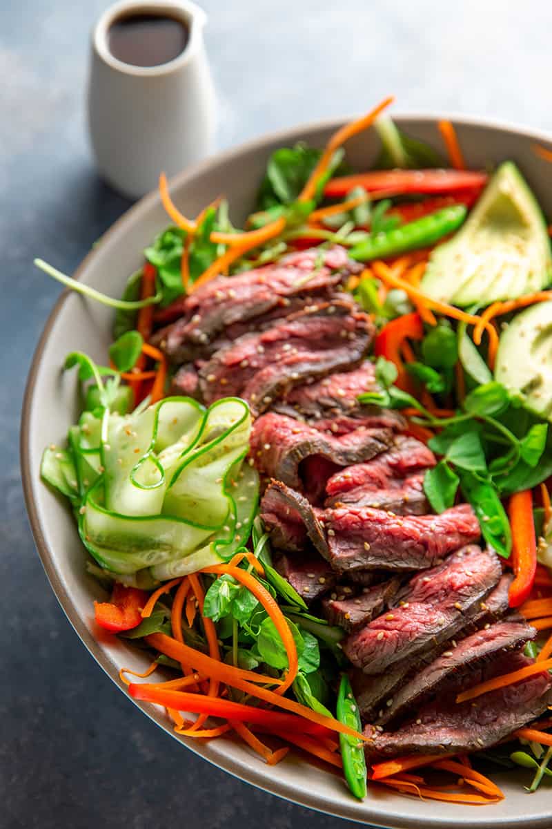 An overhead shot of Whole30 and Paleo Asian Steak Salad, a large white bowl topped with colorful vegetables and sliced seared flank steak.