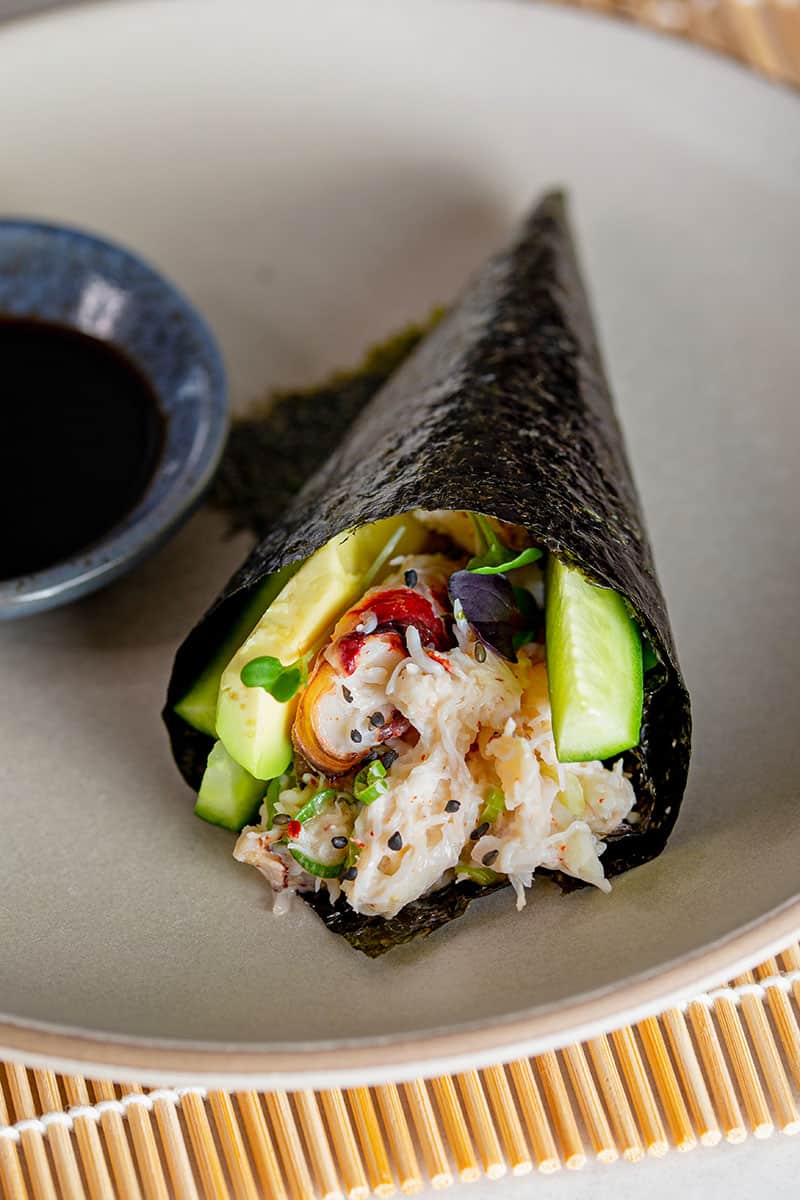 A closeup of a California hand roll on a gray plate next to a small bowl filled with coconut aminos.