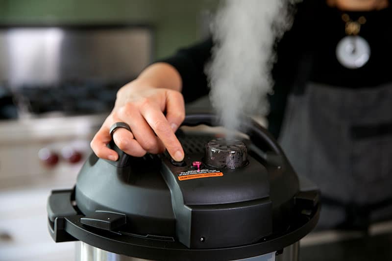 A woman is manually releasing the pressure valve on an Instant Pot and there is steam coming out of the top.
