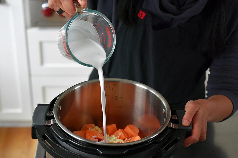 Pouring coconut milk into an Instant Pot filled with cubed sweet potatoes and minced garlic.