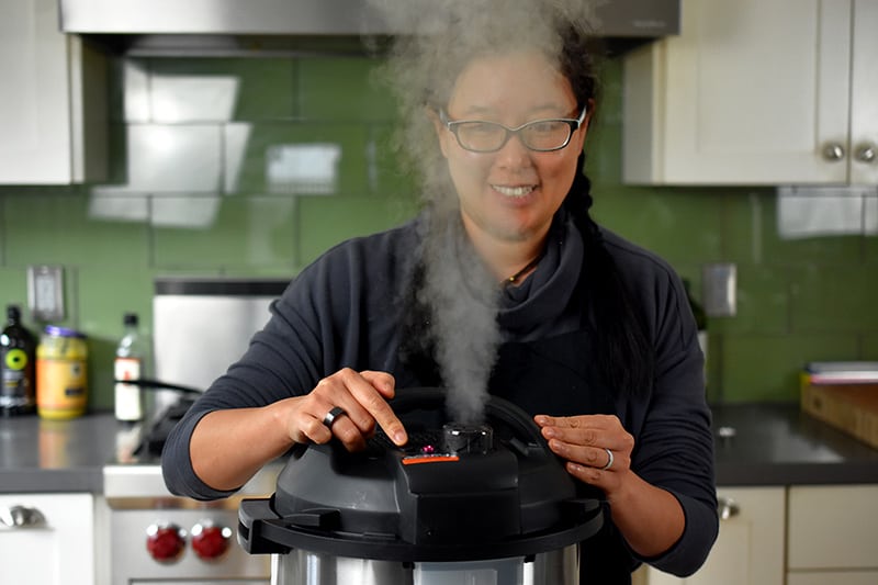 A dark haired smiling woman is manually releasing the pressure on an Instant Pot and the steam is right in front of her face.