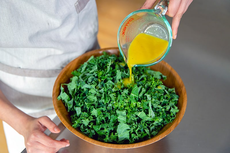 Pouring the lemon vinaigrette onto the sliced kale leaves in a big wooden bowl.