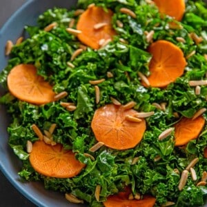 An overhead shot of two hands holding a large blue bowl filled with kale salad and persimmons and toasted almonds, a simple and delicious winter salad.