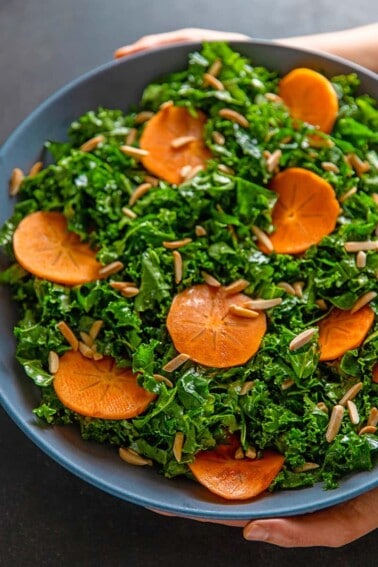 An overhead shot of two hands holding a large blue bowl filled with kale salad and persimmons and toasted almonds, a simple and delicious winter salad.
