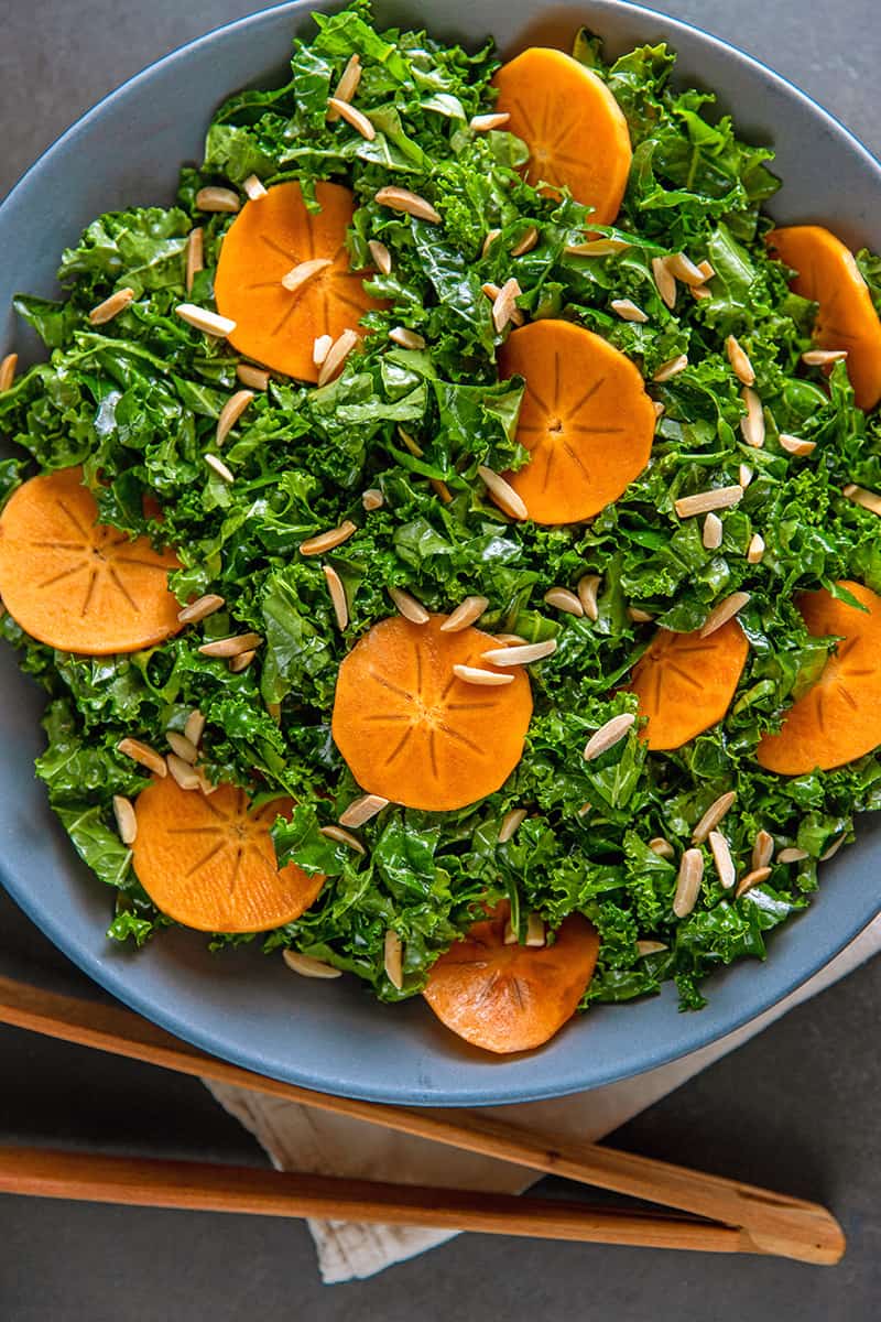 An overhead shot of a large blue bowl filled with Whole30 and vegan kale salad with persimmon next to a pair of wooden tongs.