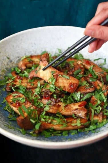 A pair of black chopsticks is picking up a piece of King trumpet mushrooms with Thai Chili peppers and lime from a light blue bowl.