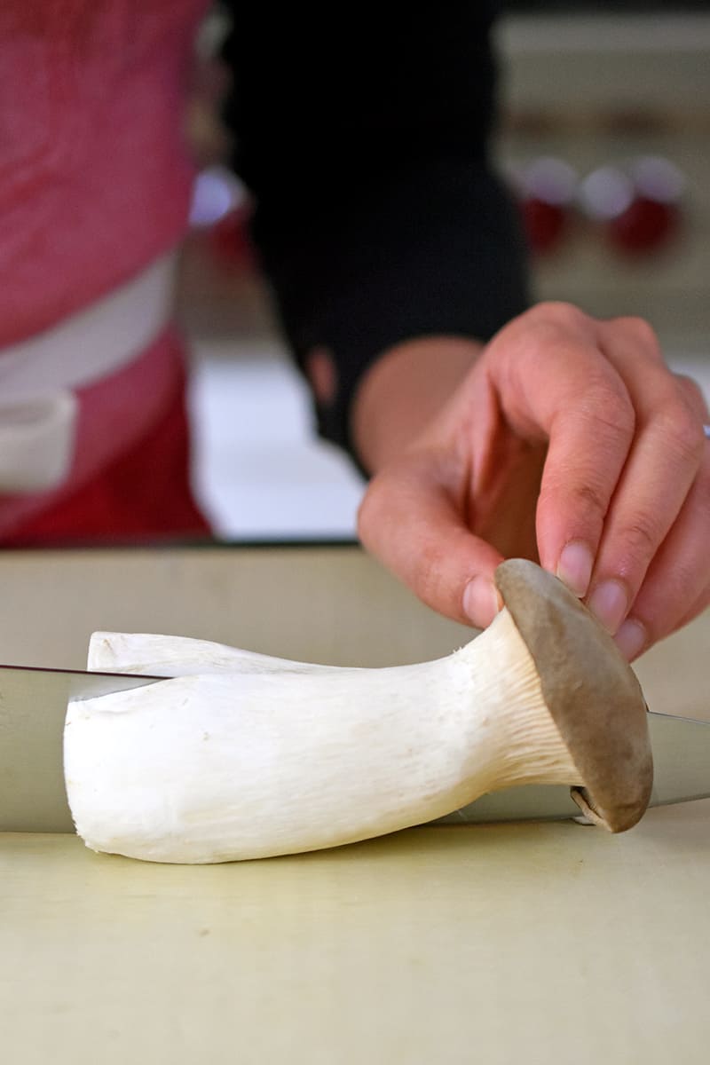 Someone using a chef's knife to slice a king oyster mushroom in half lengthwise.