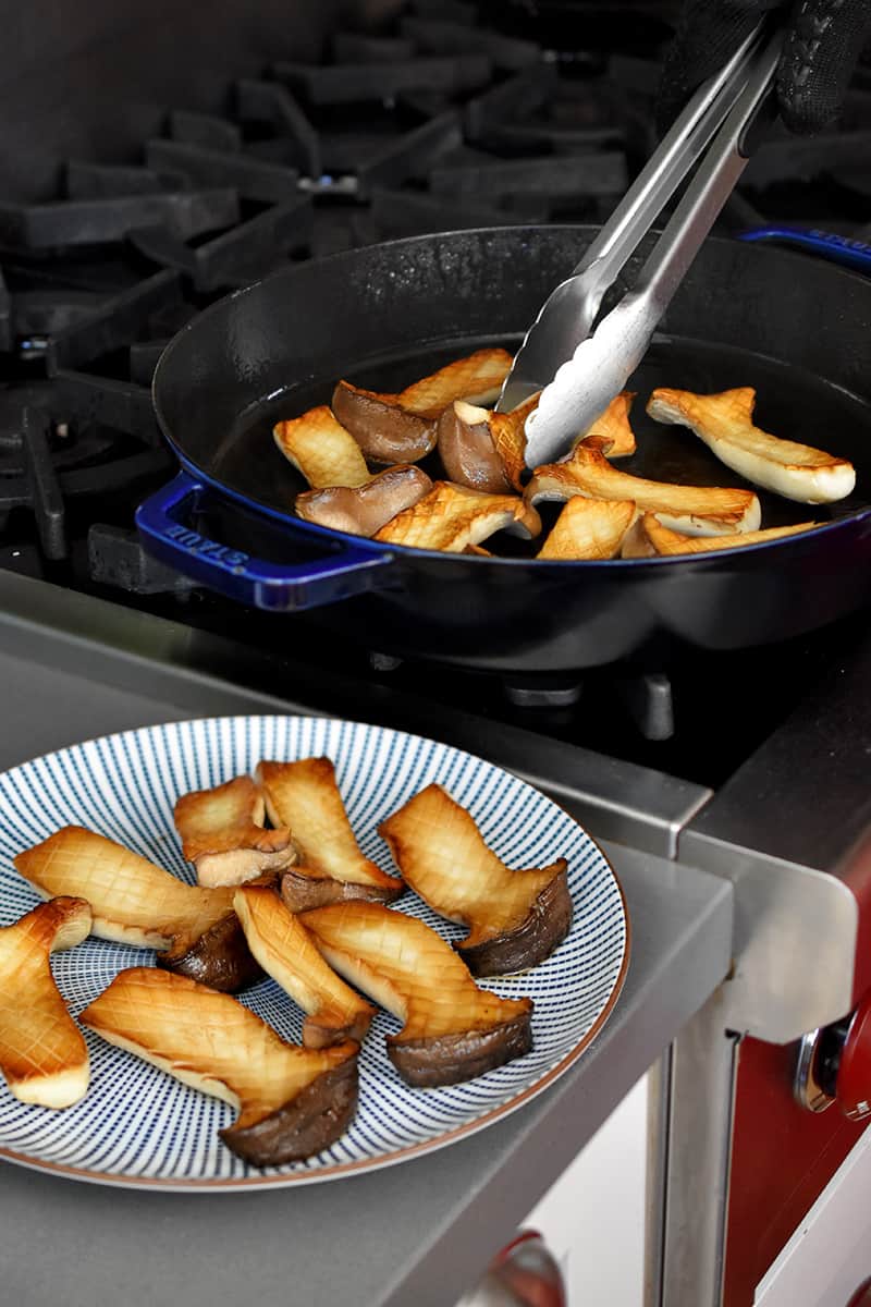 A blue and white plate has golden brown halved king oyster mushrooms and a pair of tongs are transferring the rest from a large frying pan.
