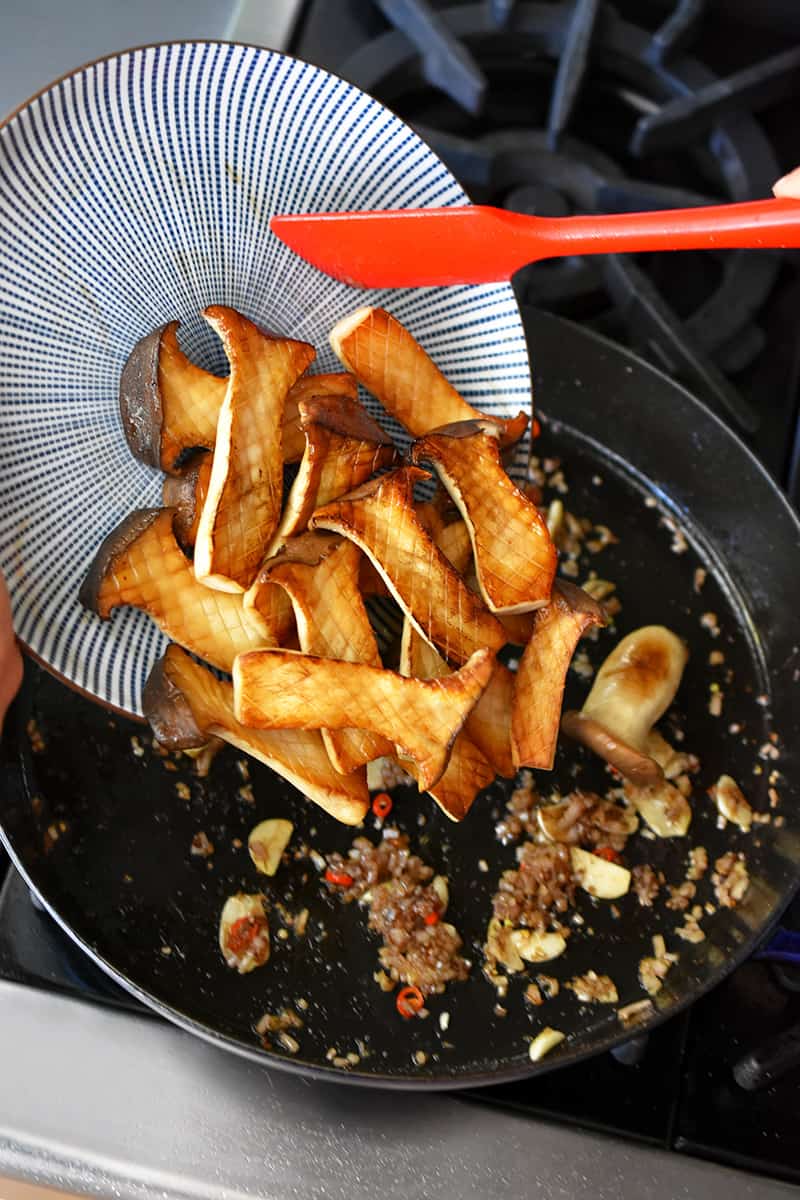 A blue and white plate filled with seared king oyster mushrooms is being added to a cast iron skillet with sautéed diced shallots, ginger, garlic, and Thai Chili peppers
