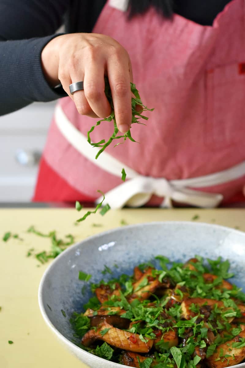 Someone in a red apron is sprinkling minced Thai basil, cilantro, and mint to a bowl filled with pan-seared king oyster mushrooms.