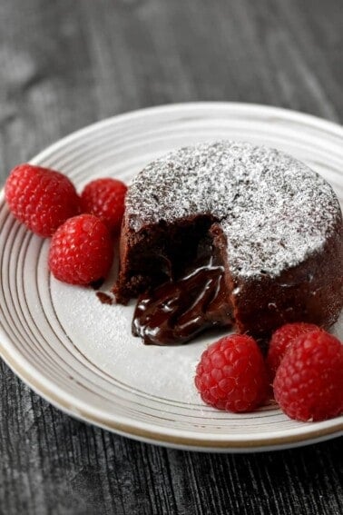 A closeup shot of a white plate with a chocolate lava cake opened to show the gooey center. There is powdered keto confectioners sugar on top and raspberries on the side.