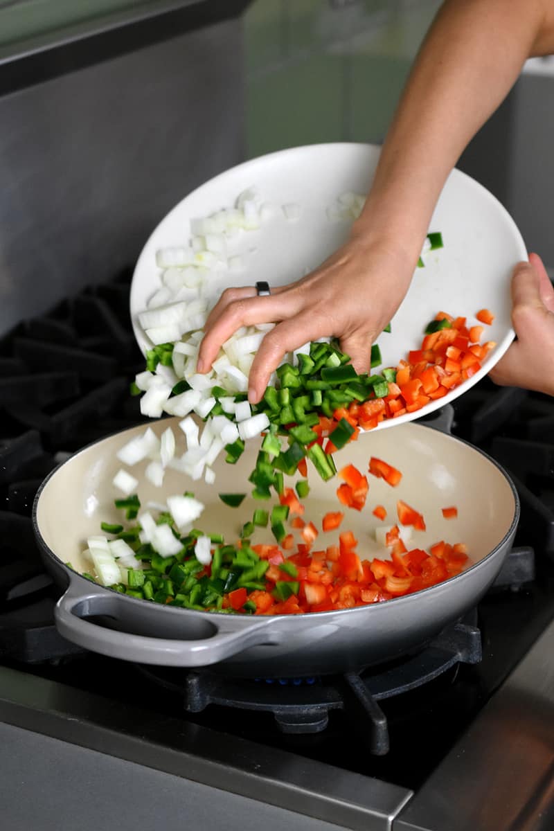 A hand is pushing diced onions, red bell peppers, and poblano peppers from a white plate into a gray enameled cast iron Le Creuset braiser.