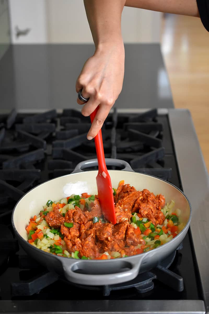 A hand is holding a red silicone spatula and it is breaking up the chorizo in the pan with diced onions and peppers.