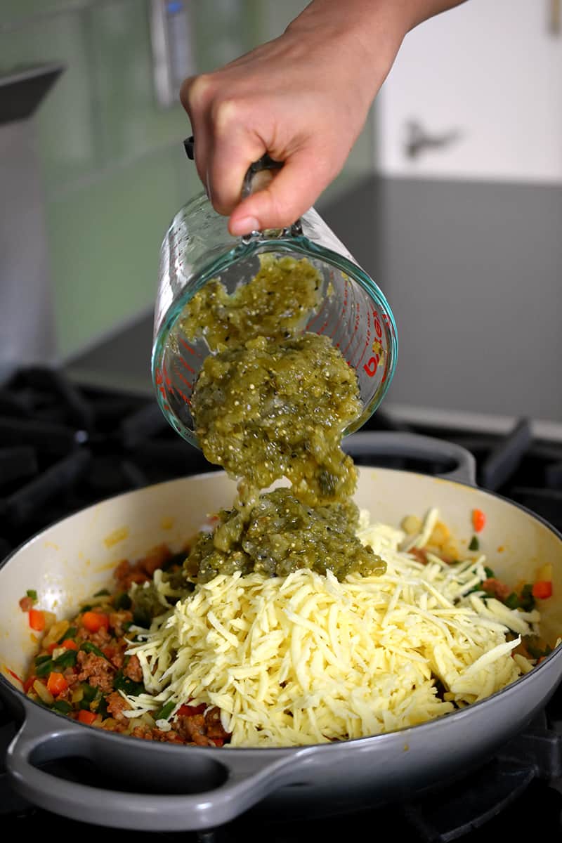 A hand pouring roasted tomatillo salsa into a pan filled with shredded white sweet potatoes, chorizo, peppers, and onions.
