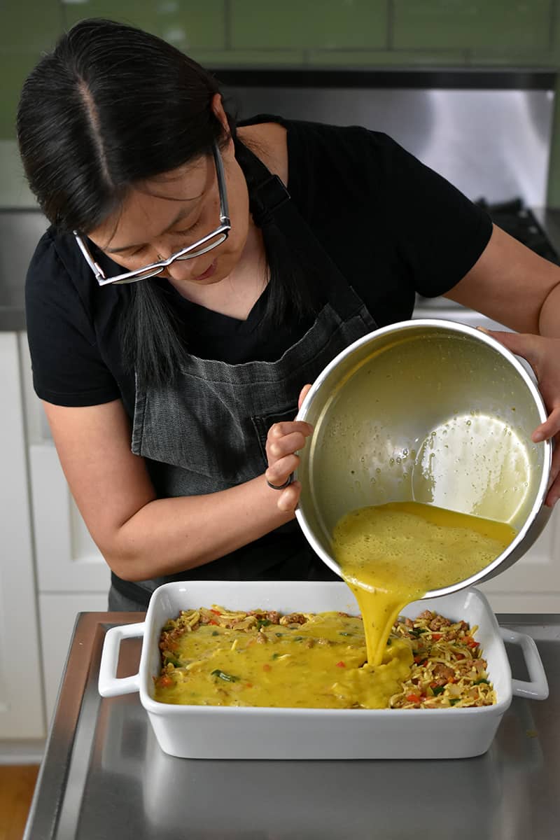 An Asian woman in glasses is pouring a big bowl of whisked eggs into a casserole pan to make Mexican Breakfast Casserole.