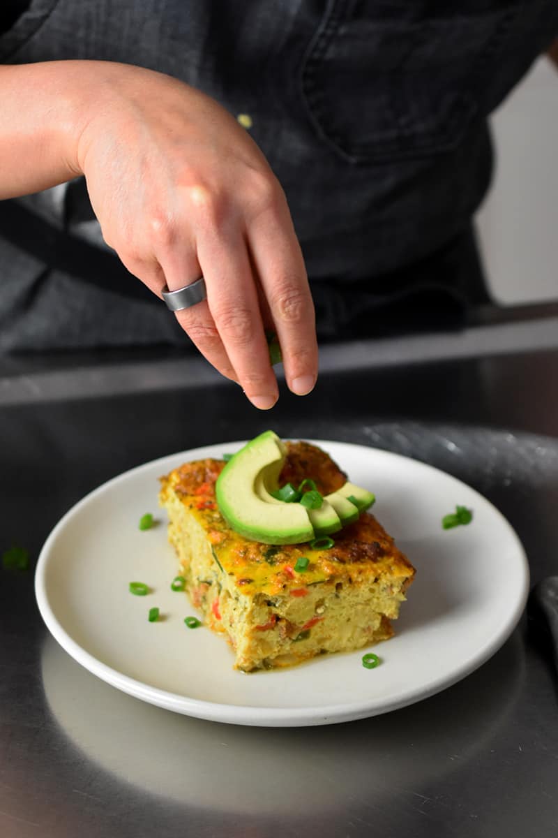 A hand is sprinkling sliced scallions on a slice of Mexican breakfast casserole with avocado slices on top