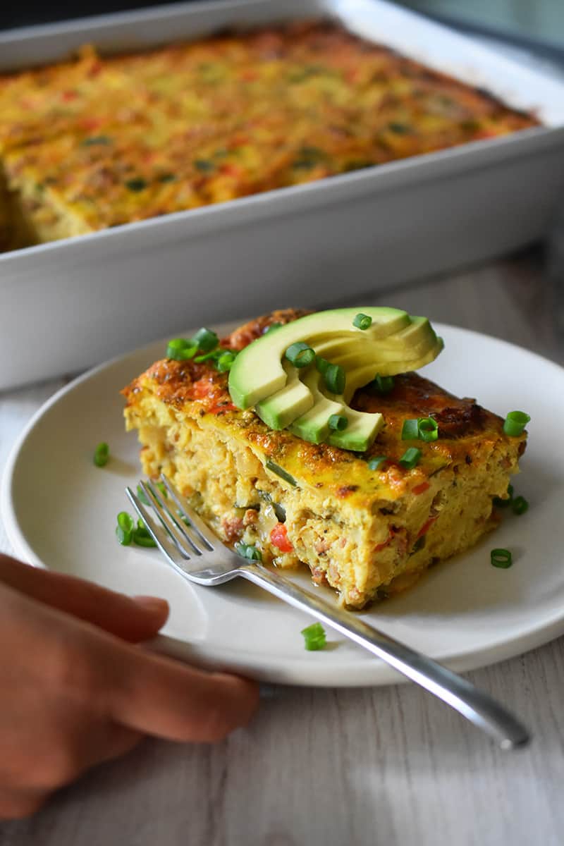A hand is grabbing a plate with a slice of Mexican breakfast casserole.