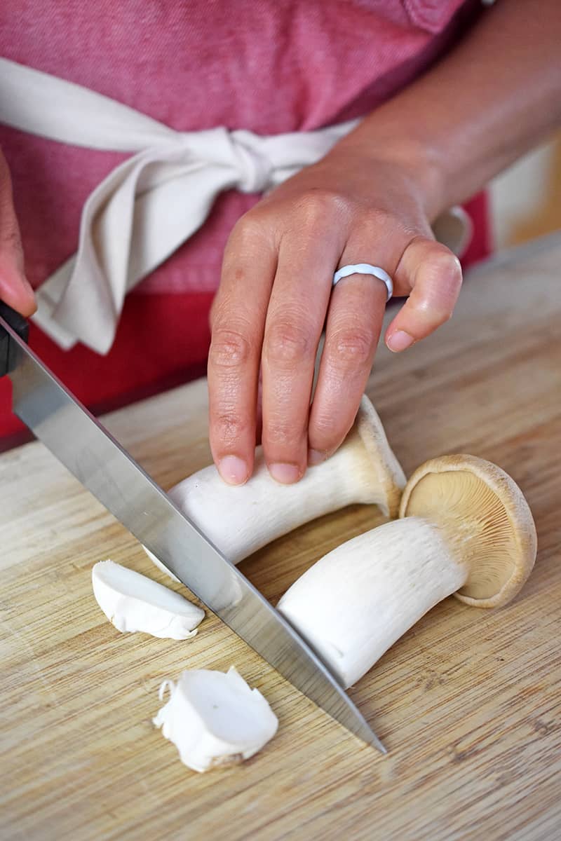 Someone in a red apron is cutting the bottoms off of two king oyster mushrooms.