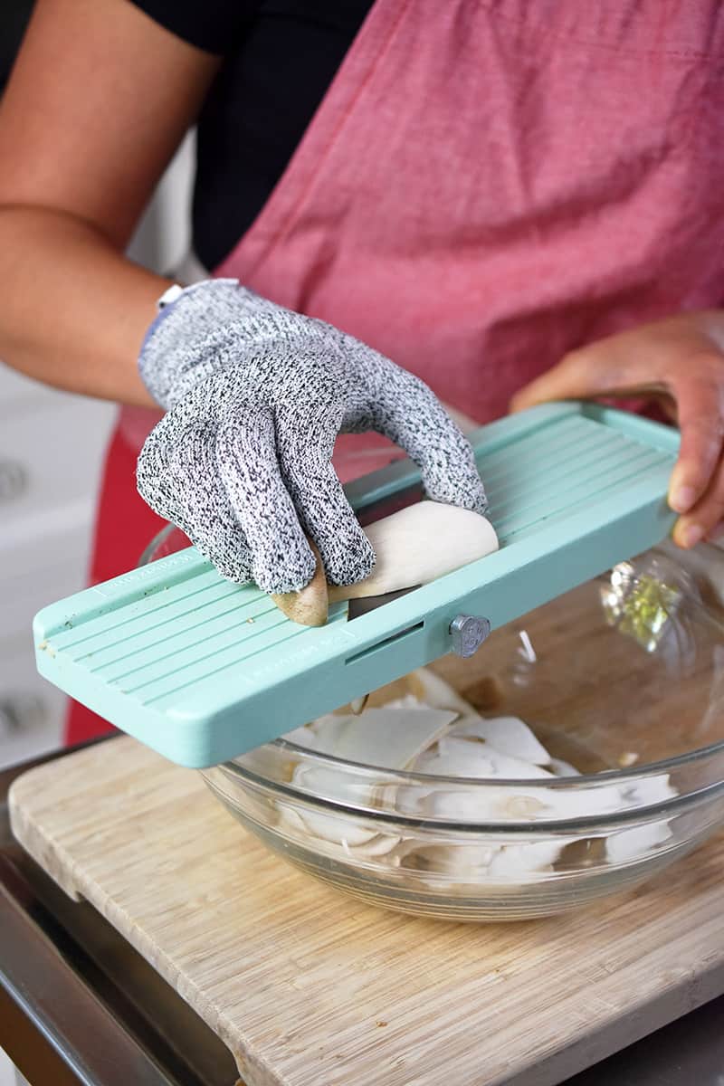 Someone wearing cut-resistant gloves is using a Japanese mandolin to shave king oyster mushrooms.