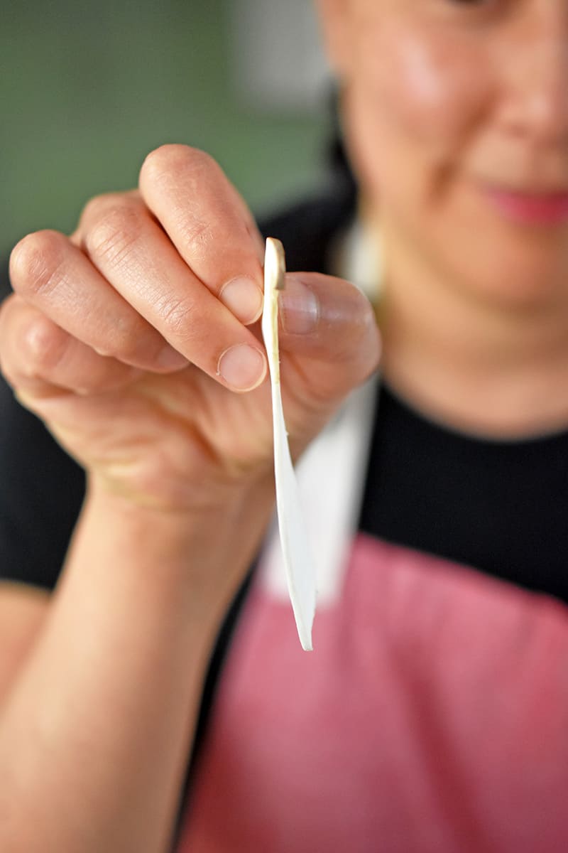 A woman in a red apron is holding up a paper thin slice of king oyster mushroom.