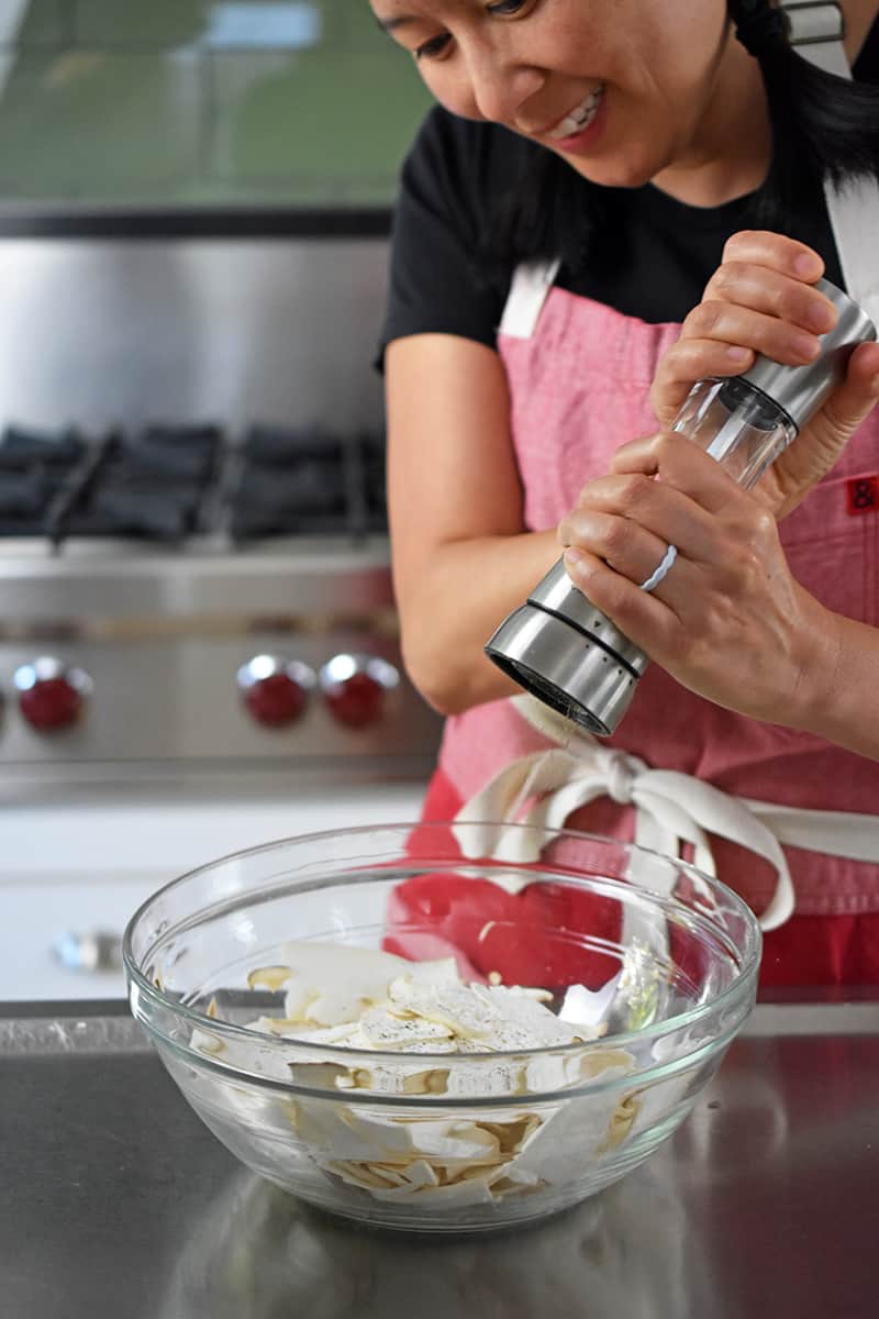 A woman in a red apron is adding freshly cracked black pepper into a large bowl filled with shaved king oyster mushrooms.