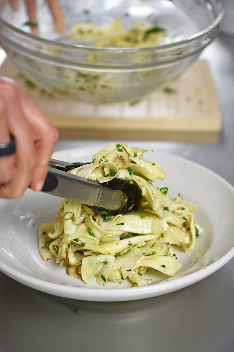 A pair of tongs is transferring the mushroom salad to a serving platter