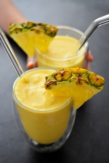 An overhead shot of two clear glasses filled with spicy turmeric smoothie. The cups are garnished with a [ineapple wedge and there are glass straws in the cups.