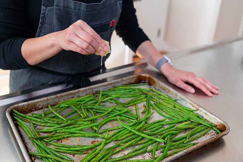 Squeezing a lemon wedge onto a pan of roasted green beans with garlic.