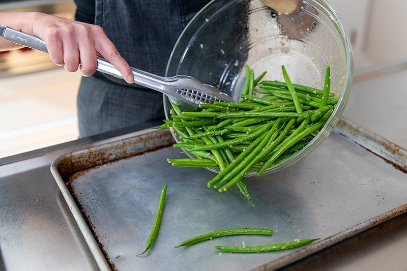 Transferring the marinated green beans to a rimmed baking sheet with a pair of tongs.
