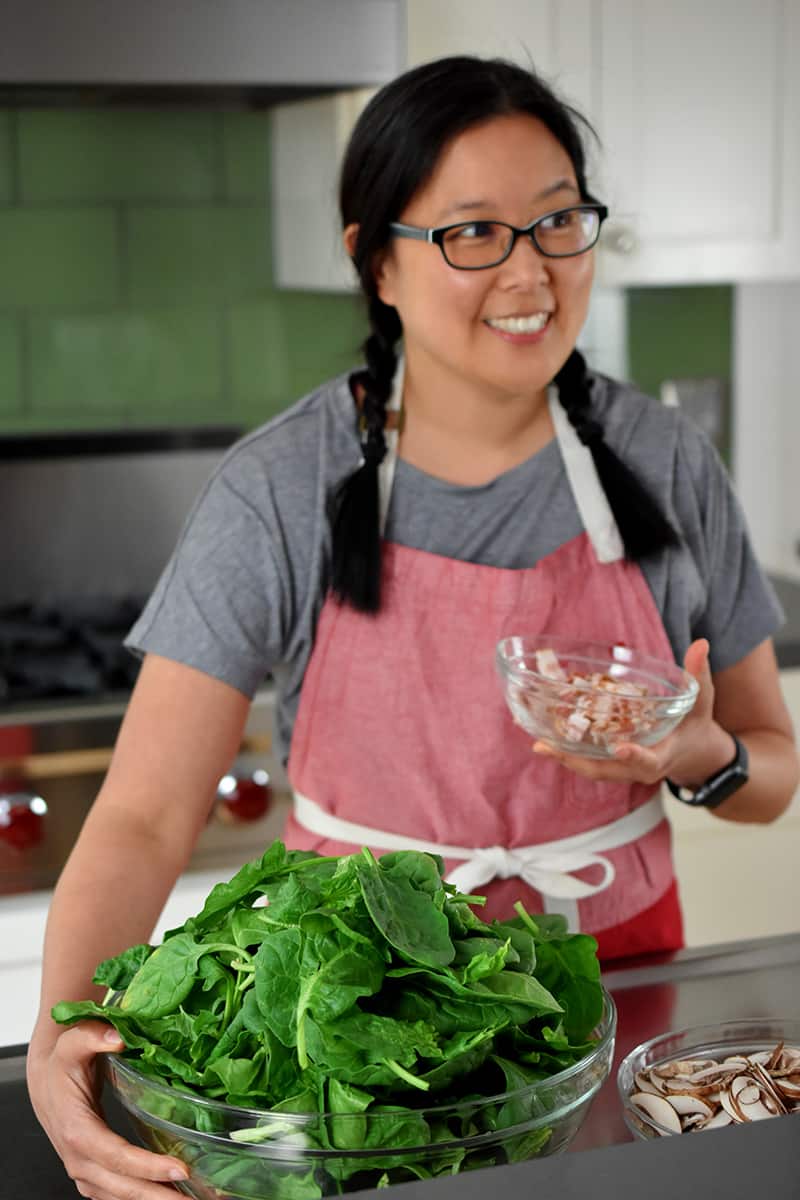 An Asian woman in a red apron is holding a large bowl of raw spinach and sliced bacon.