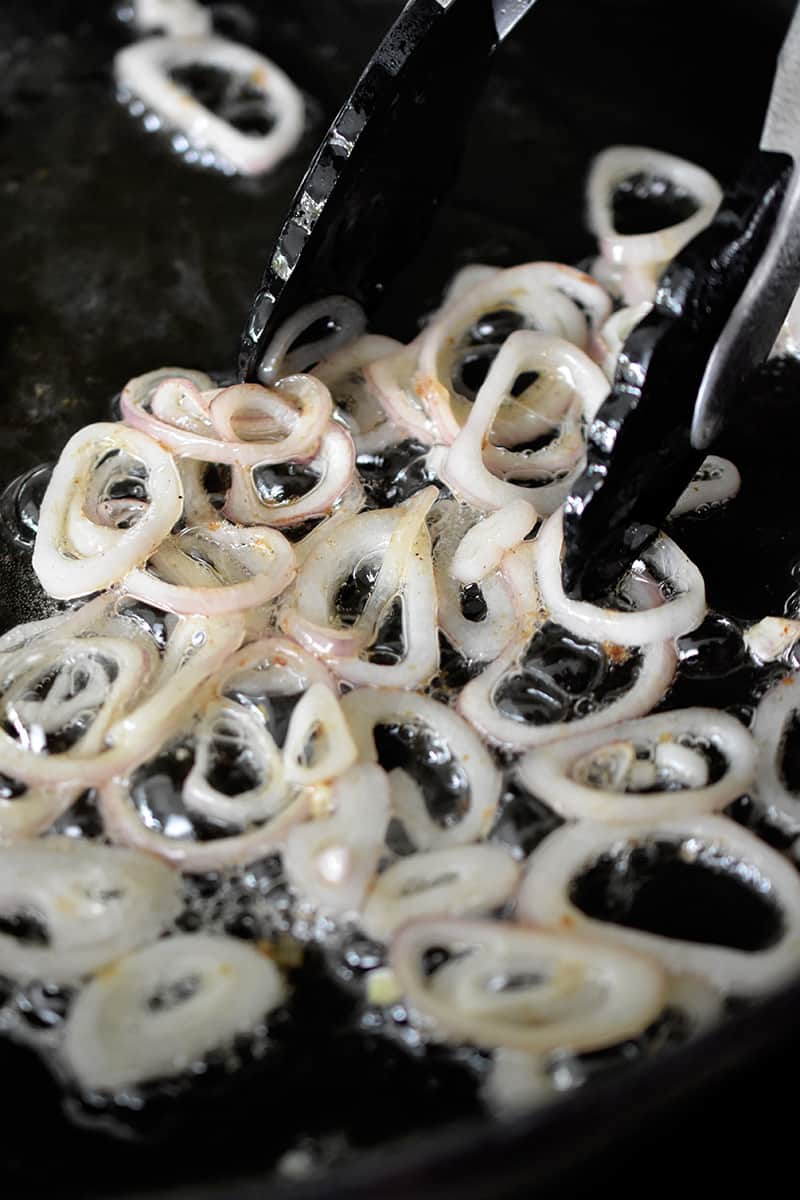 A pair of tongs are pushing thin shallot rings in a cast iron skillet.