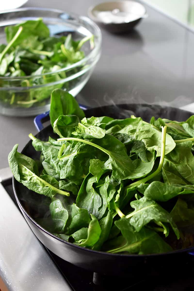 There is a bowl with spinach in it next to a large skillet filled with spinach.