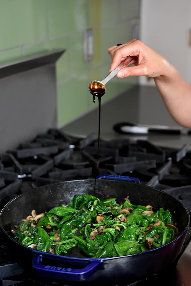 A hand is asking a spoonful of aged balsamic to a pan filled with spinach, mushrooms, and shallots.