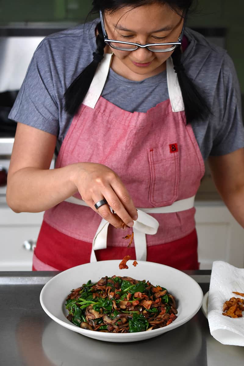 A woman in a red apron is sprinkling crispy bacon bits on top of a bowl filled with sautéed spinach and mushrooms.