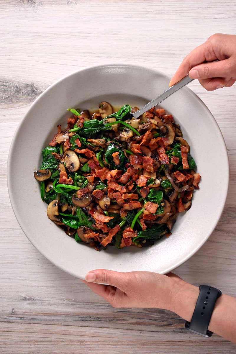 To hands are holding a cream colored bowl filled with sautéed spinach with bacon and mushrooms.