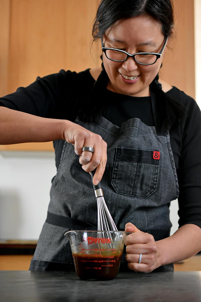 A smiling Asian woman is whisking All-Purpose Stir-Fry Sauce with other seasonings in a 2 cup Pyrex measuring cup
