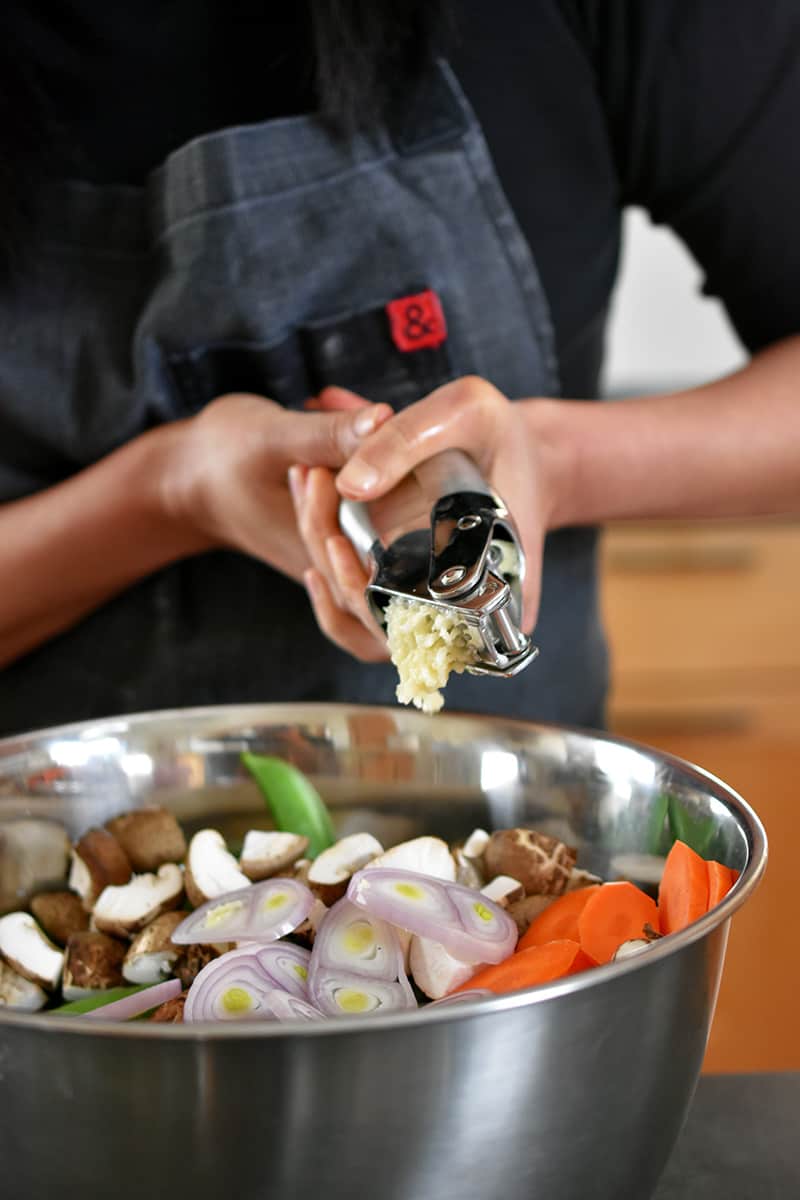 Someone in a gray apron is squeezing a garlic press with two hands and adding the minced garlic to a big silver bowl filled with chopped vegetables