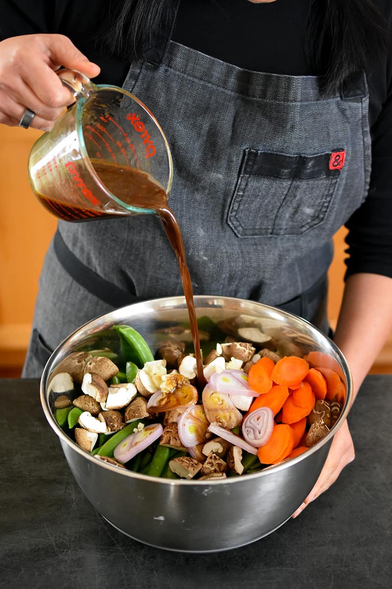 A person in a gray apron is pouring a brown sauce into a large metal bowl filled with chopped carrots, sugar snap peas, shallots, shiitake mushrooms, and minced garlic.