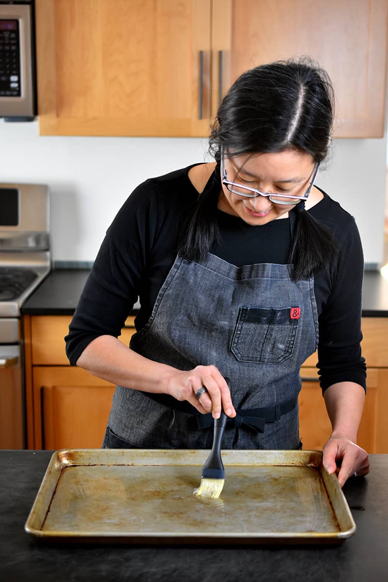 A brunette Asian woman is brushing avocado oil on a rimmed baking sheet.