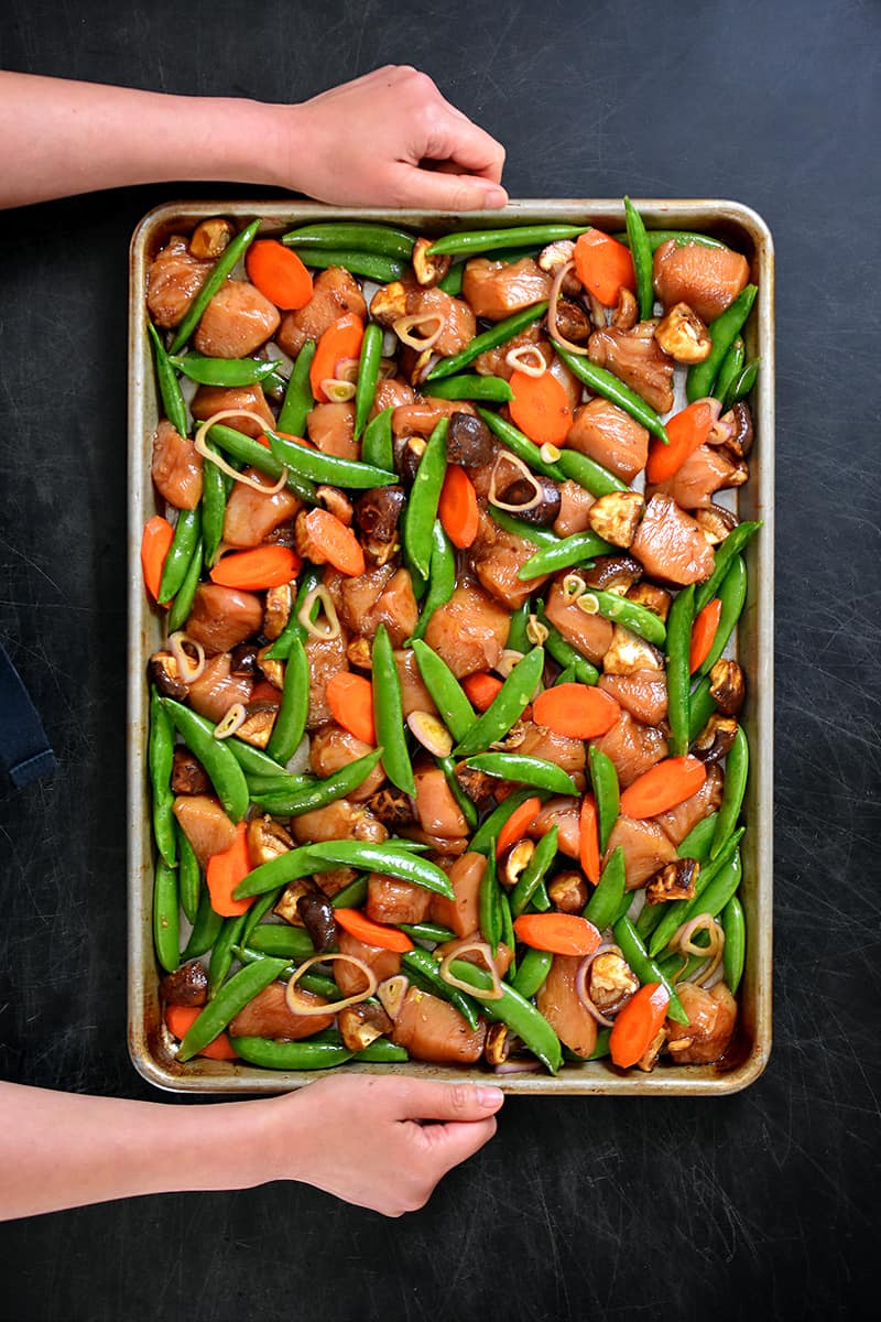 An overhead shot of a sheet pan filled with uncooked stir-fry chicken and vegetables in a single layer.