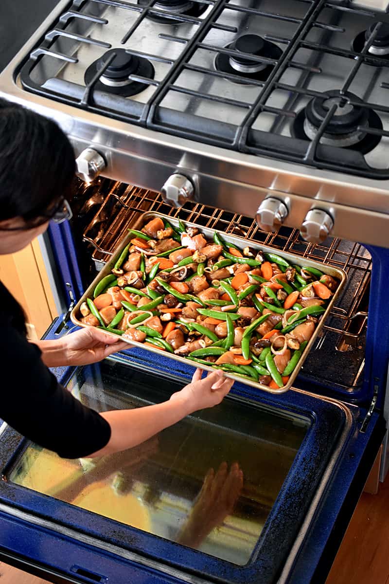 An Asian woman is bending down to place a sheet pan filled with chicken and vegetables into an open oven.