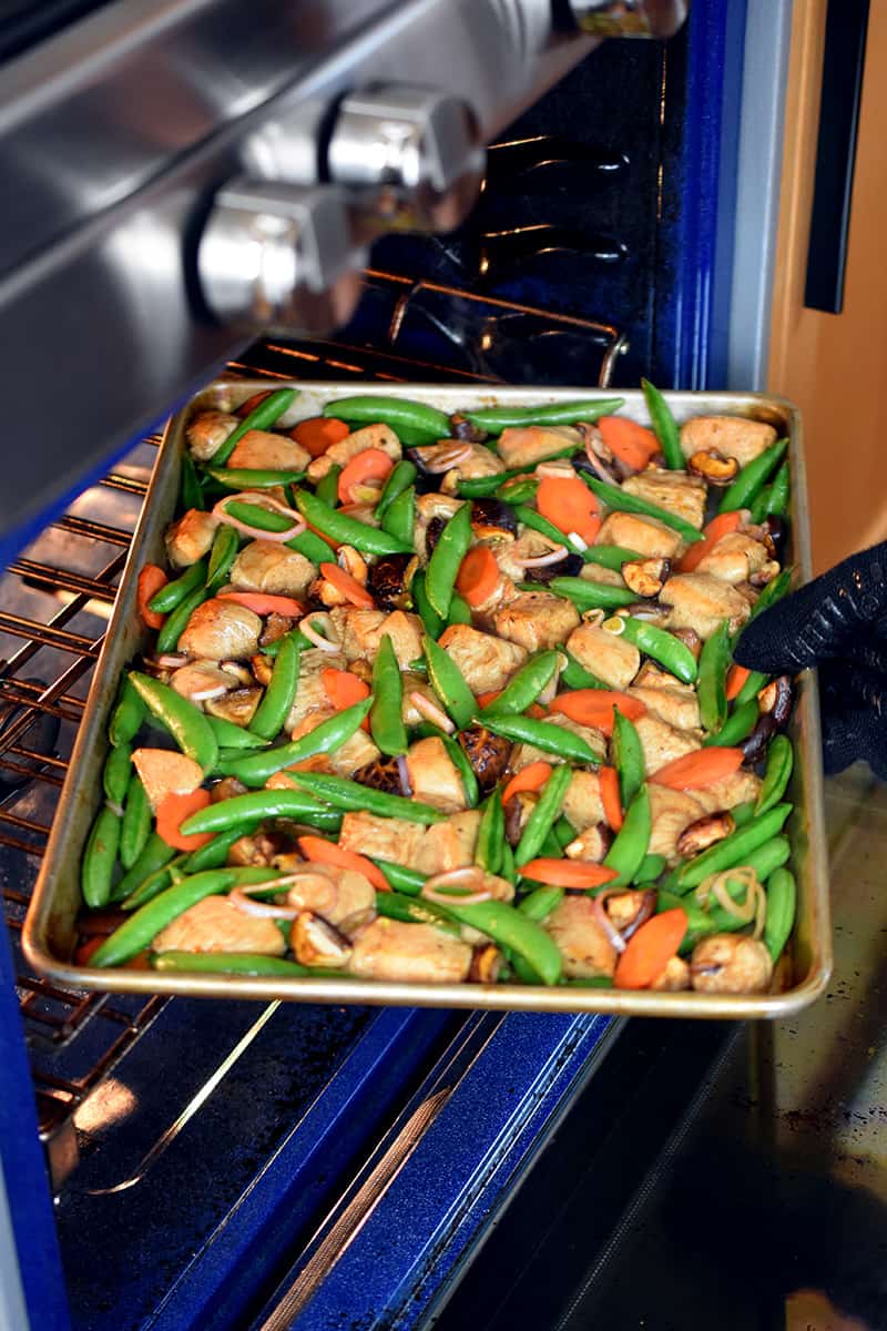 A gloved hand is rotating a sheet pan chicken and vegetable dinner at the midway point while it is baking.