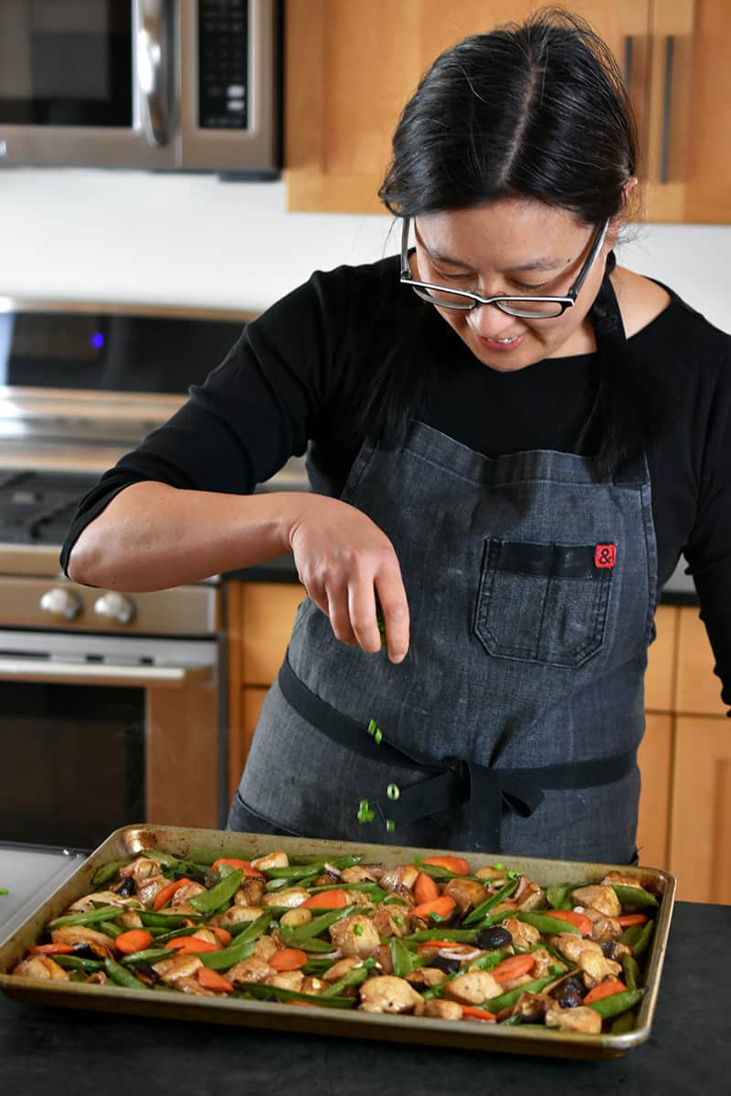 An Asian woman is sprinkling sliced scallions on a sheet pan filled with stir-fry chicken and vegetables.