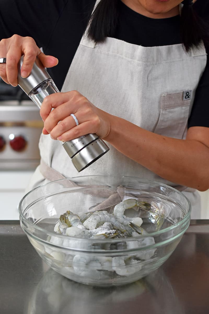 A person in a beige apron is adding freshly cracked black pepper to a bowl of peeled raw shrimp.