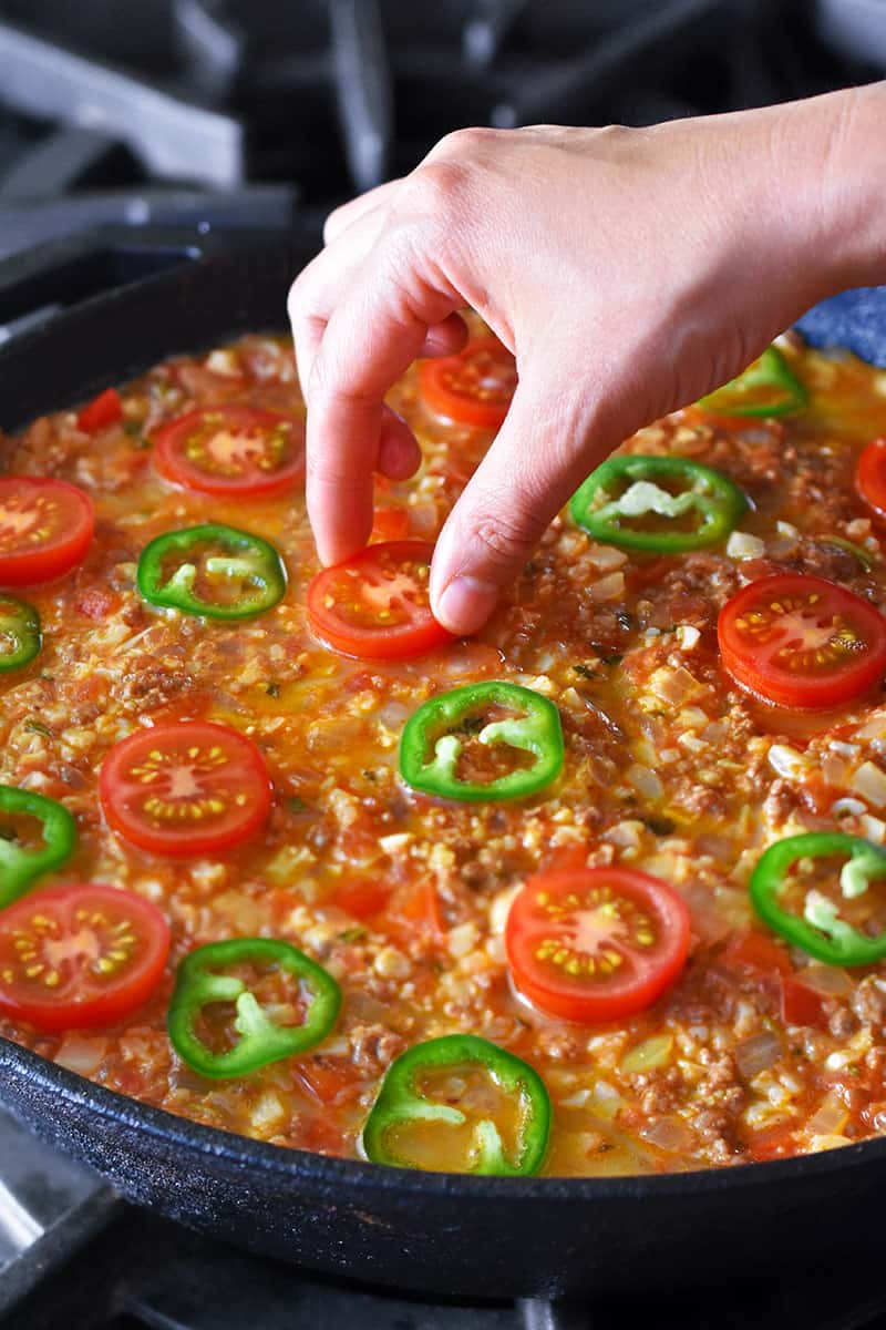 Adding halved cherry tomatoes and sliced jalapeño peppers on top of paleo taco casserole with ground beef and cauliflower rice.