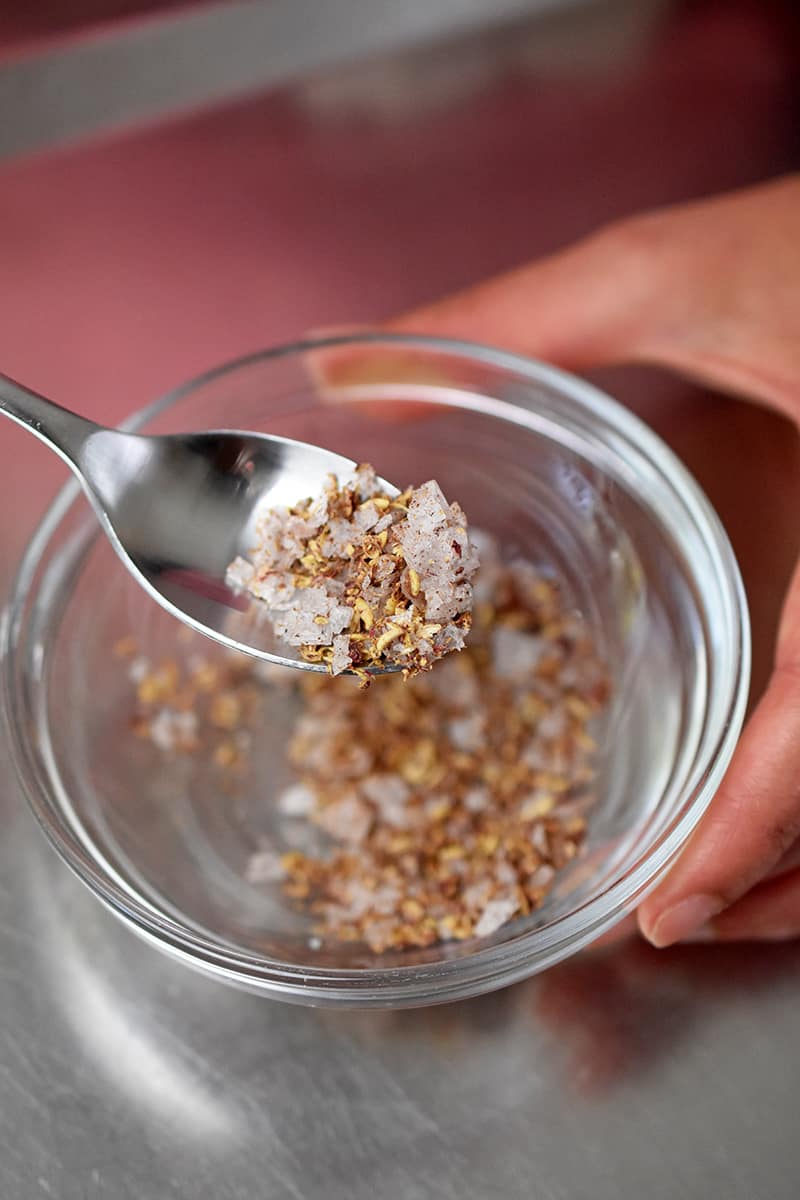 An overhead shot of a spoon mixing crushed Sichuan pepper corns with flaky sea salt in a small bowl.